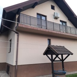 Elewacje z klasą - Dwukondygnacyjny dom z balkonem i klimatyzacją, beżowa elewacja z brązowym cokołem, studnia na pierwszym planie, widok z dołu.