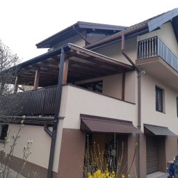 Elewacje z klasą - Dwukondygnacyjny dom z beżowo-brązową elewacją, drewnianą pergolą na balkonie i metalowymi zadaszeniami nad garażem. W tle drzewo i fragment budynku sąsiada.