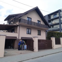 Elewacje z klasą - Dwupiętrowy dom z balkonem i brązową bramą, obok którego stoi grupa robotników z materiałami izolacyjnymi. W tle widoczny nowoczesny budynek.