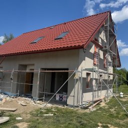 Elewacje z klasą - Dom jednorodzinny w trakcie tynkowania elewacji, widoczne rusztowania, czerwona dachówka i błękitne niebo. Prace wykończeniowe w toku.