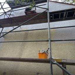 Elewacje z klasą - Dwóch pracowników na rusztowaniu maluje elewację domu na żółto i biało. Widoczne narzędzia malarskie i wiaderka z farbą. Częściowo pomalowana ściana z wyraźną fakturą.
