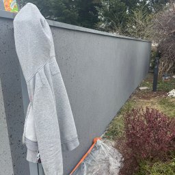Elewacje z klasą - Świeżo pomalowany, szary mur ogrodzeniowy z widoczną fakturą, z bliska. Na murze wisi szara bluza z kapturem, a u dołu widoczne zabezpieczenia malarskie.