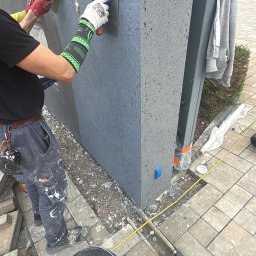 Elewacje z klasą - Praca przy elewacji: dwóch mężczyzn nakłada szary tynk na ścianę budynku. Widoczne narzędzia, wiadro z zaprawą i fragment posesji. Jeden z pracowników ma zielone rękawice.