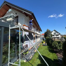 Elewacje z klasą - Praca na rusztowaniu: mężczyzna w niebieskiej bluzie aplikuje jasny tynk na elewację domu z ciemnymi oknami. Widoczne narzędzia i przygotowane podłoże.