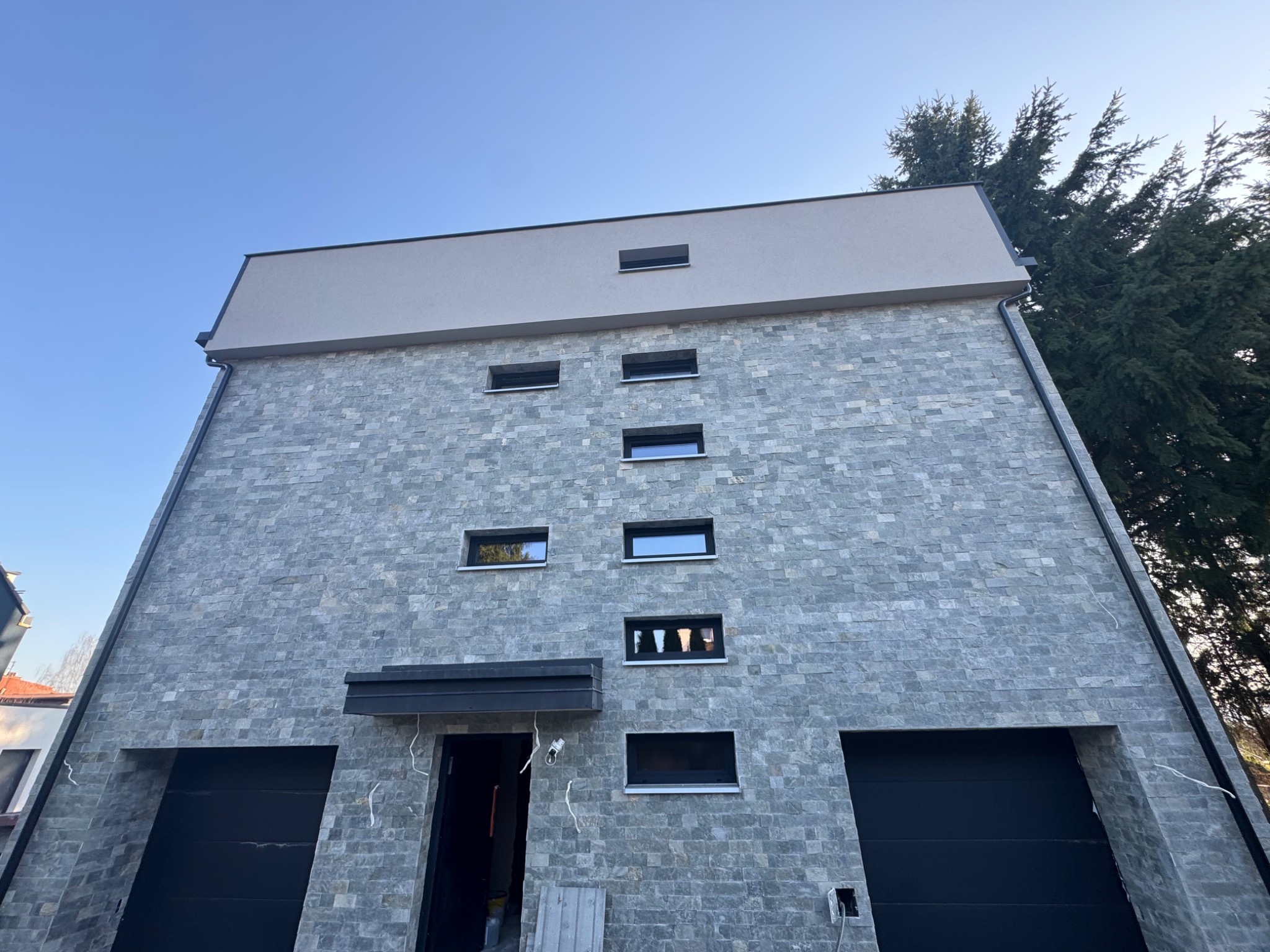 Nowoczesny dom z elewacją z szarego kamienia, czarne okna i bramy garażowe. Widok z dołu, błękitne niebo w tle. Częściowo widoczne przewody elektryczne.