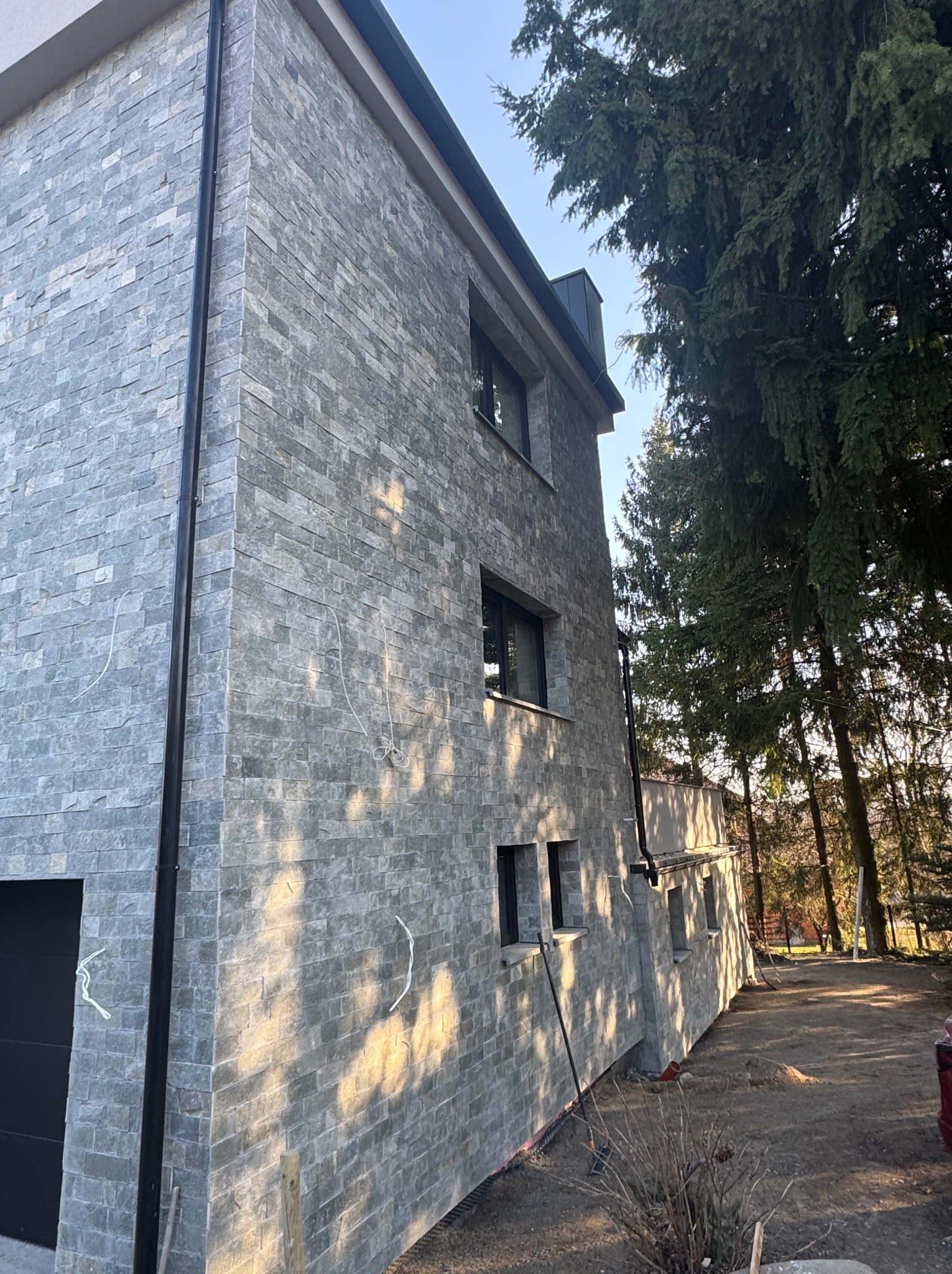 Elewacja domu wykończona szarą, kamienną okładziną, widok z dołu, z fragmentem garażu i drzewami w tle, w słoneczny dzień. Widoczne przewody elektryczne.