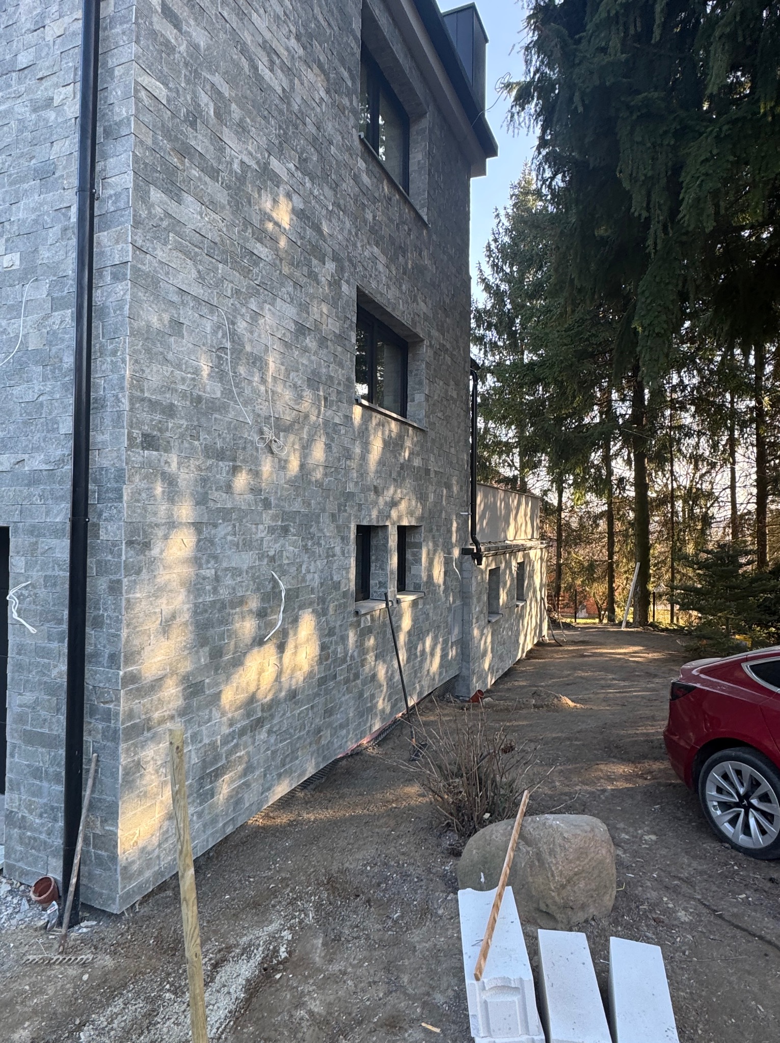 Elewacja budynku wykończona szarą, kamienną okładziną. Widoczne okna, fragment dachu, drzewa w tle i czerwony samochód. Częściowo widoczne materiały budowlane.
