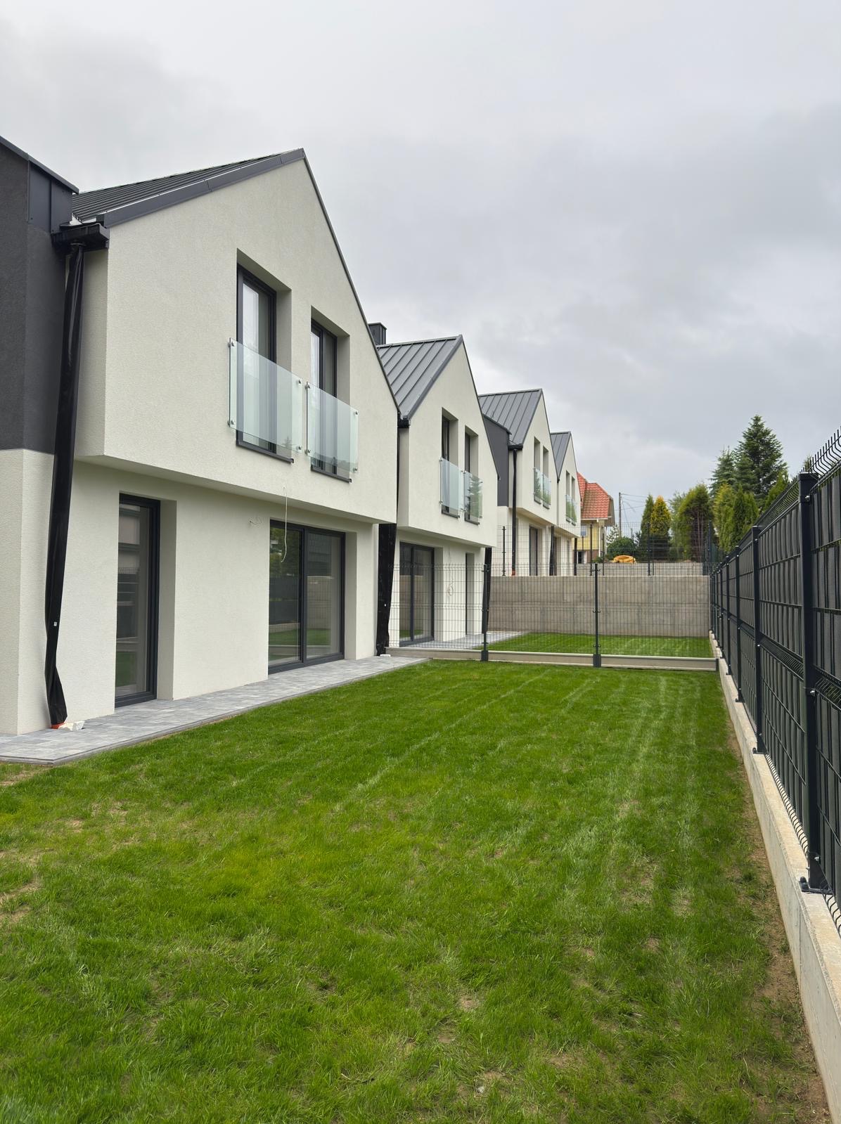 Rząd nowoczesnych domów z białymi elewacjami i szarymi dachami, widok z perspektywy trawnika oddzielonego czarnym ogrodzeniem panelowym. Pochmurne niebo.