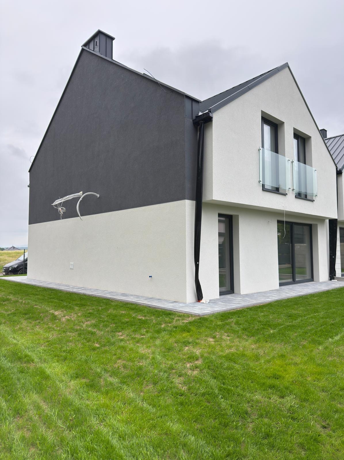 Dwukolorowa elewacja nowoczesnego domu z balkonem i szklanymi balustradami. Widoczne elementy wykończeniowe i trawnik. Ujęcie z boku, w pochmurny dzień.