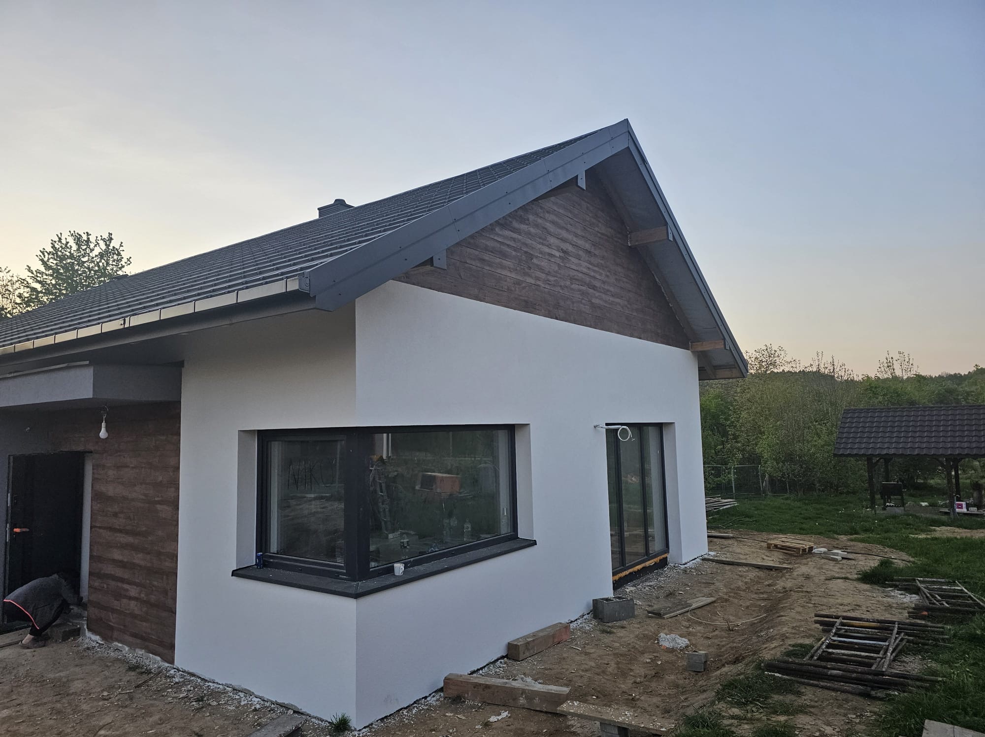 Nowoczesny dom jednorodzinny z białą elewacją i drewnianymi akcentami, widoczny z boku, w trakcie prac wykończeniowych na zewnątrz. Uwagę zwraca duże okno.