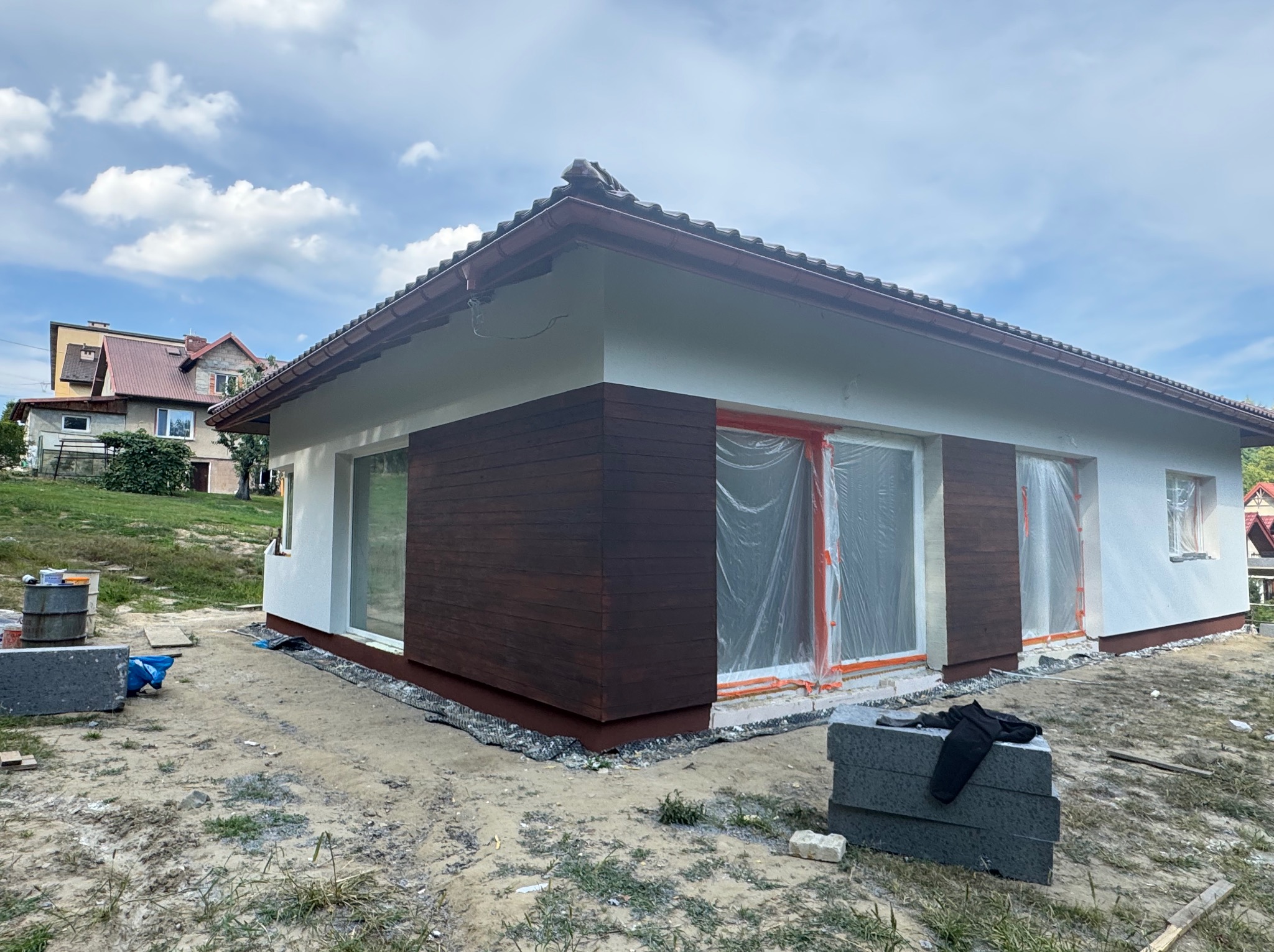 Dom jednorodzinny z częściowo wykończoną elewacją: białe ściany i brązowe panele drewnopodobne. Okna zabezpieczone folią malarską. Widoczne materiały budowlane.