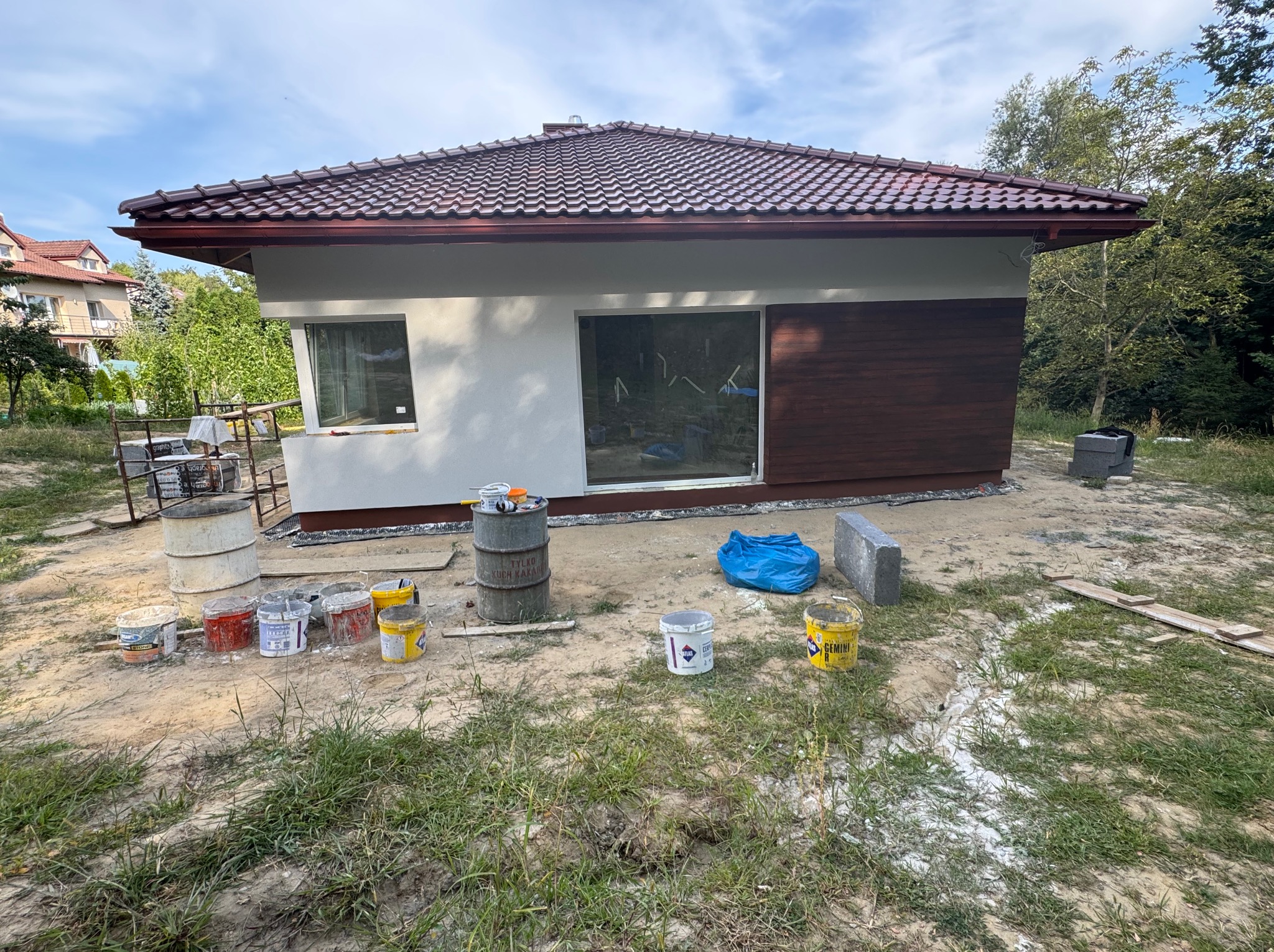 Świeżo pomalowany budynek z brązowym dachem i drewnianą elewacją. Widoczne puszki z farbą i narzędzia malarskie. Prace wykończeniowe w toku.