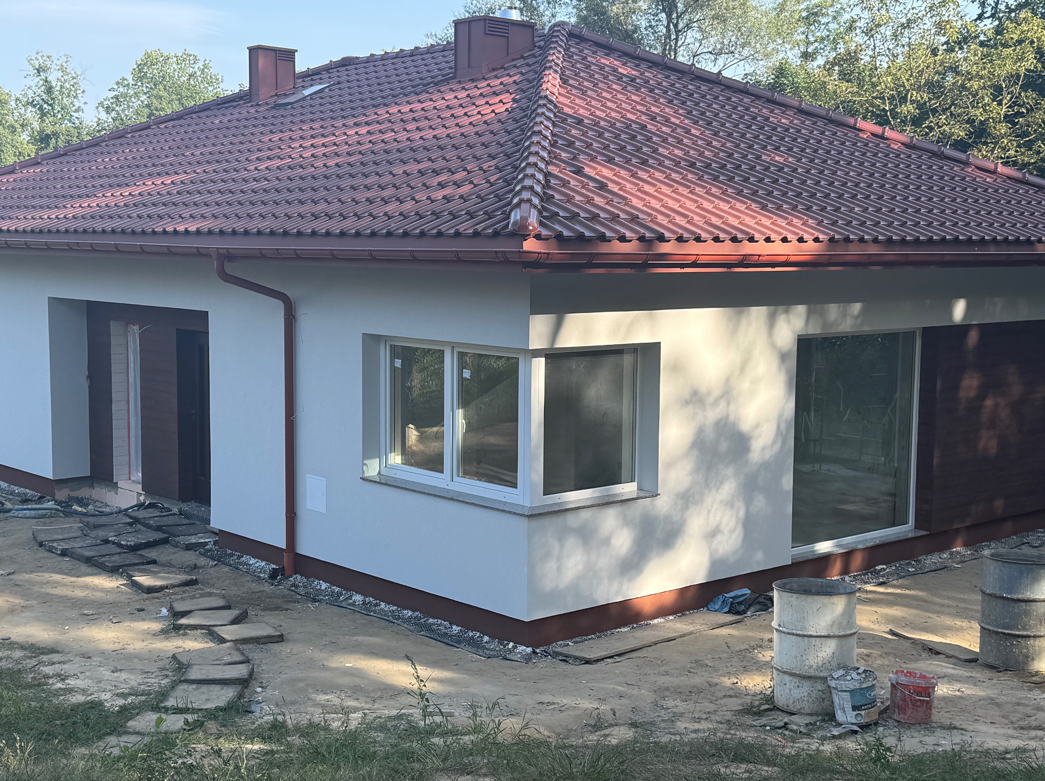 Parterowy dom z czerwoną dachówką i jasną elewacją w trakcie prac wykończeniowych. Widoczne narzędzia i materiały budowlane. Ujęcie w słoneczny dzień.