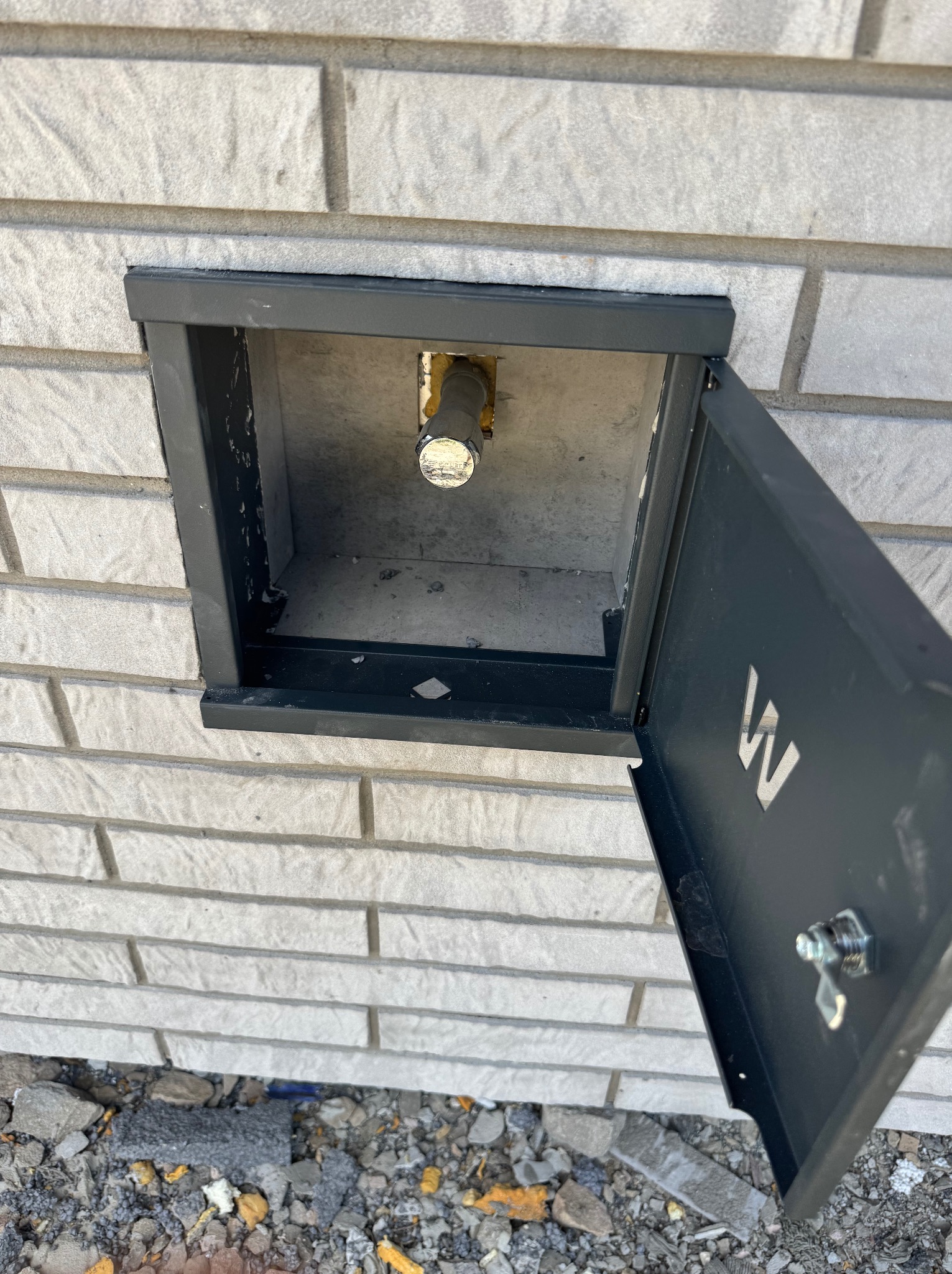 Biała ceglana ściana z czarną, otwartą skrzynką rewizyjną, wewnątrz której widoczny jest zawór. Otwarta klapka z wyciętą literą 'W'.