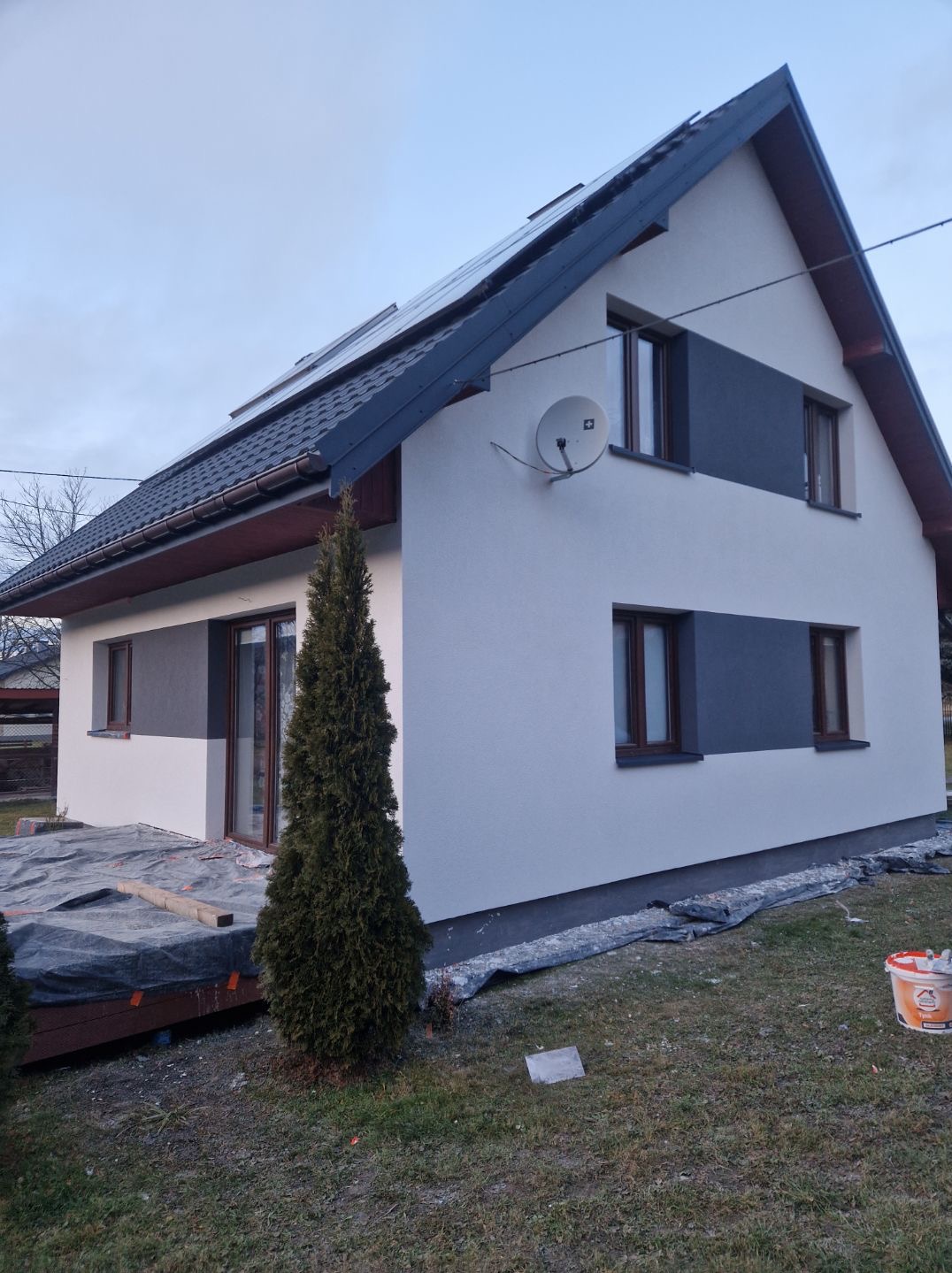 Dwukondygnacyjny dom z nową, dwukolorową elewacją (biało-szara). Widoczne okna z brązowymi ramami, antena satelitarna i częściowo zakryty taras. Ujęcie z poziomu gruntu.