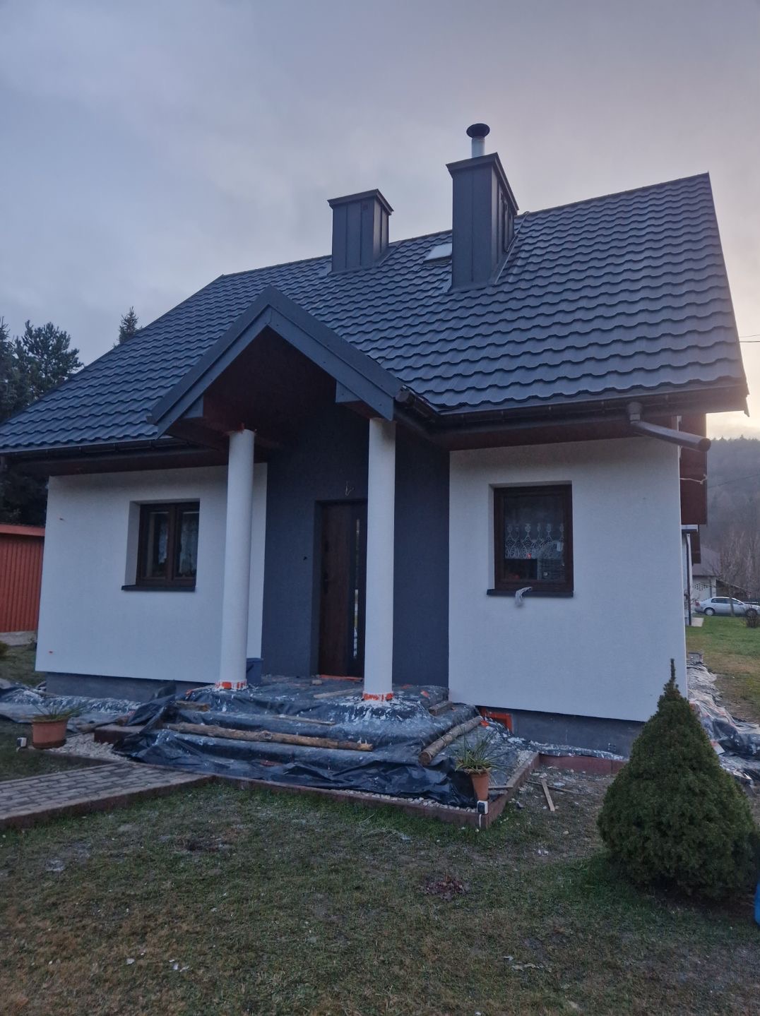 Dom jednorodzinny po malowaniu elewacji na biało i szaro, widoczne zabezpieczenia przed zabrudzeniem. Ujęcie z poziomu trawnika, pochmurne niebo w tle.