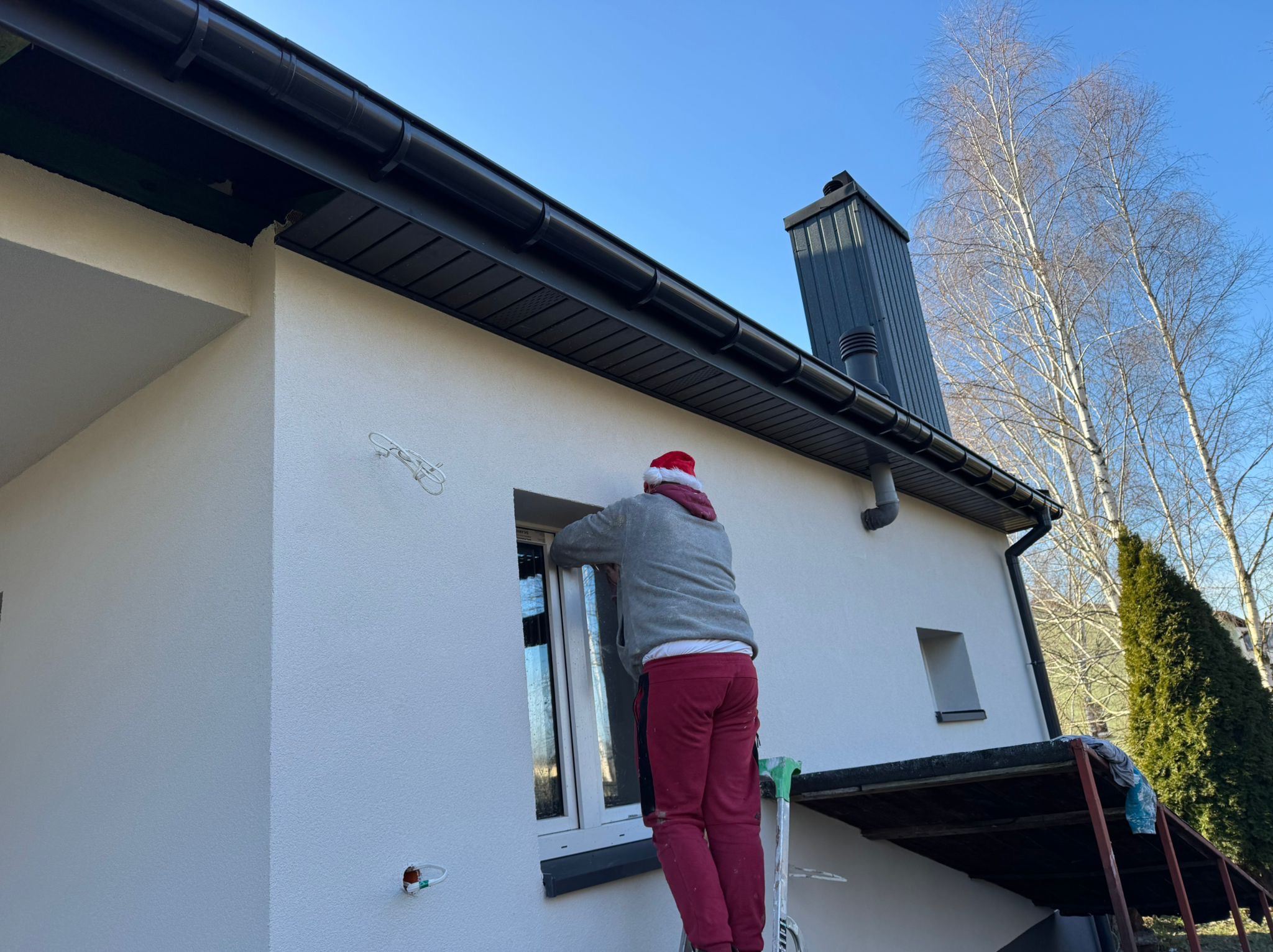 Pracownik w czapce Mikołaja na drabinie, uszczelnia okno w świeżo otynkowanej, białej elewacji domu z czarnym dachem i rynnami. Błękitne niebo w tle.