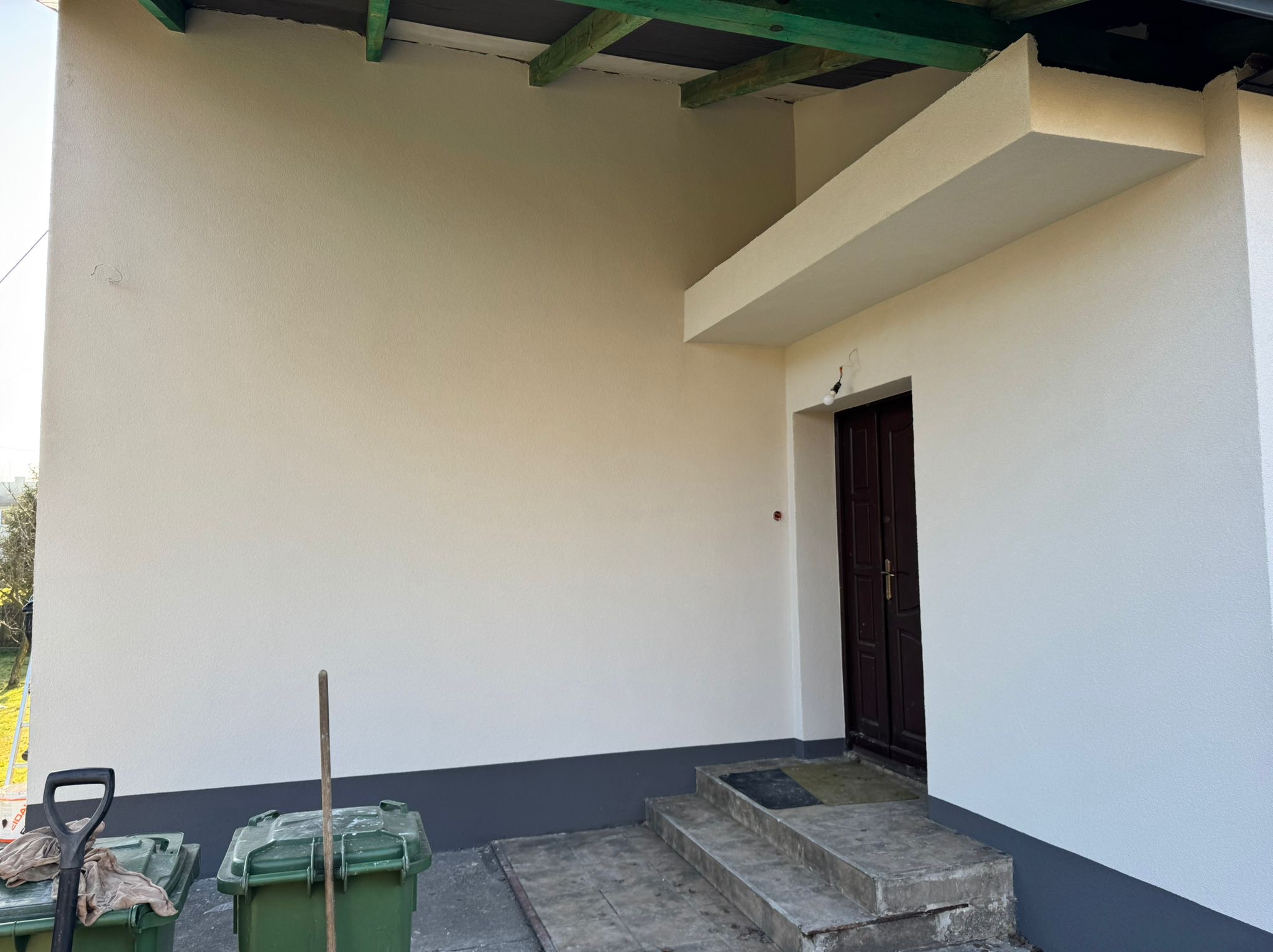 Jasna elewacja domu po renowacji, z ciemnoszarą podmurówką i wejściem. Widoczne narzędzia i pojemniki na odpady. Świeżo pomalowana ściana z widoczną fakturą.