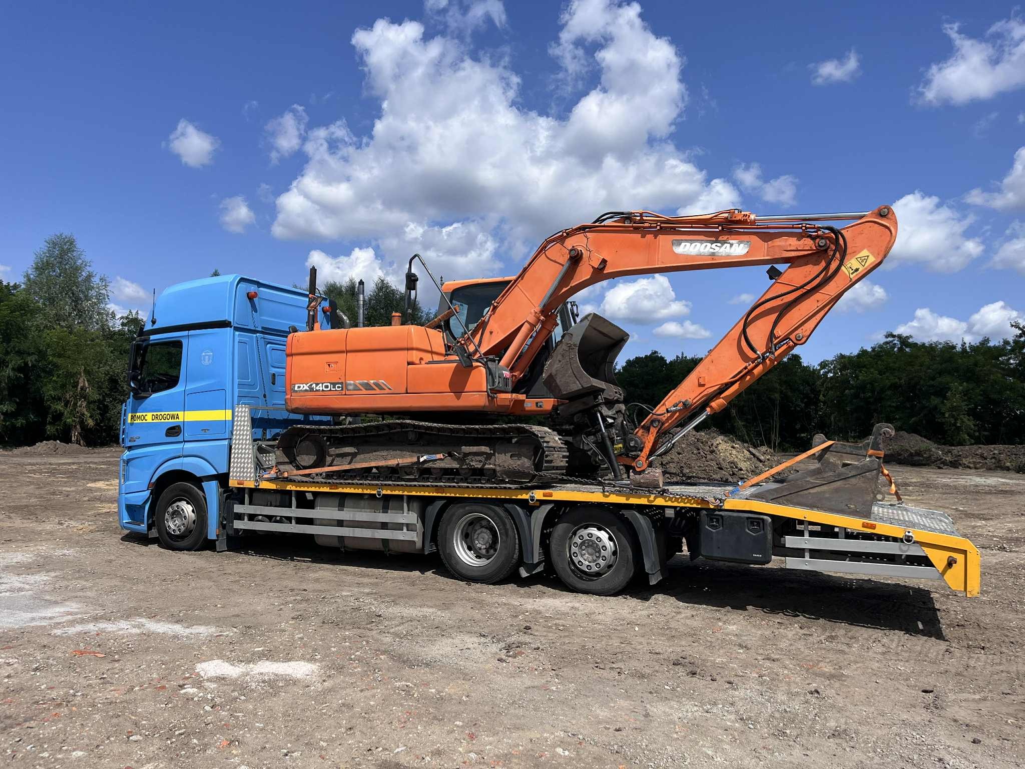 Niebieska laweta z pomarańczową koparką Doosan DX140LC na platformie, widok z boku na tle błękitnego nieba i zieleni. Na kabinie napis 'Pomoc Drogowa'.