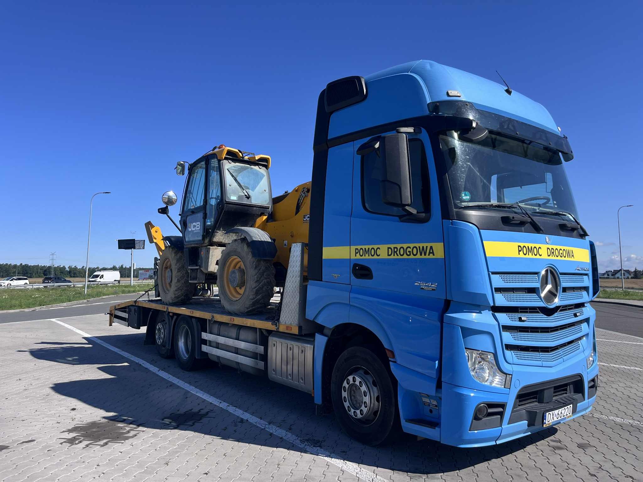 Niebieska laweta Mercedes-Benz z żółtą koparko-ładowarką JCB na platformie, zaparkowana na parkingu w słoneczny dzień. Widoczny napis 'Pomoc Drogowa'.