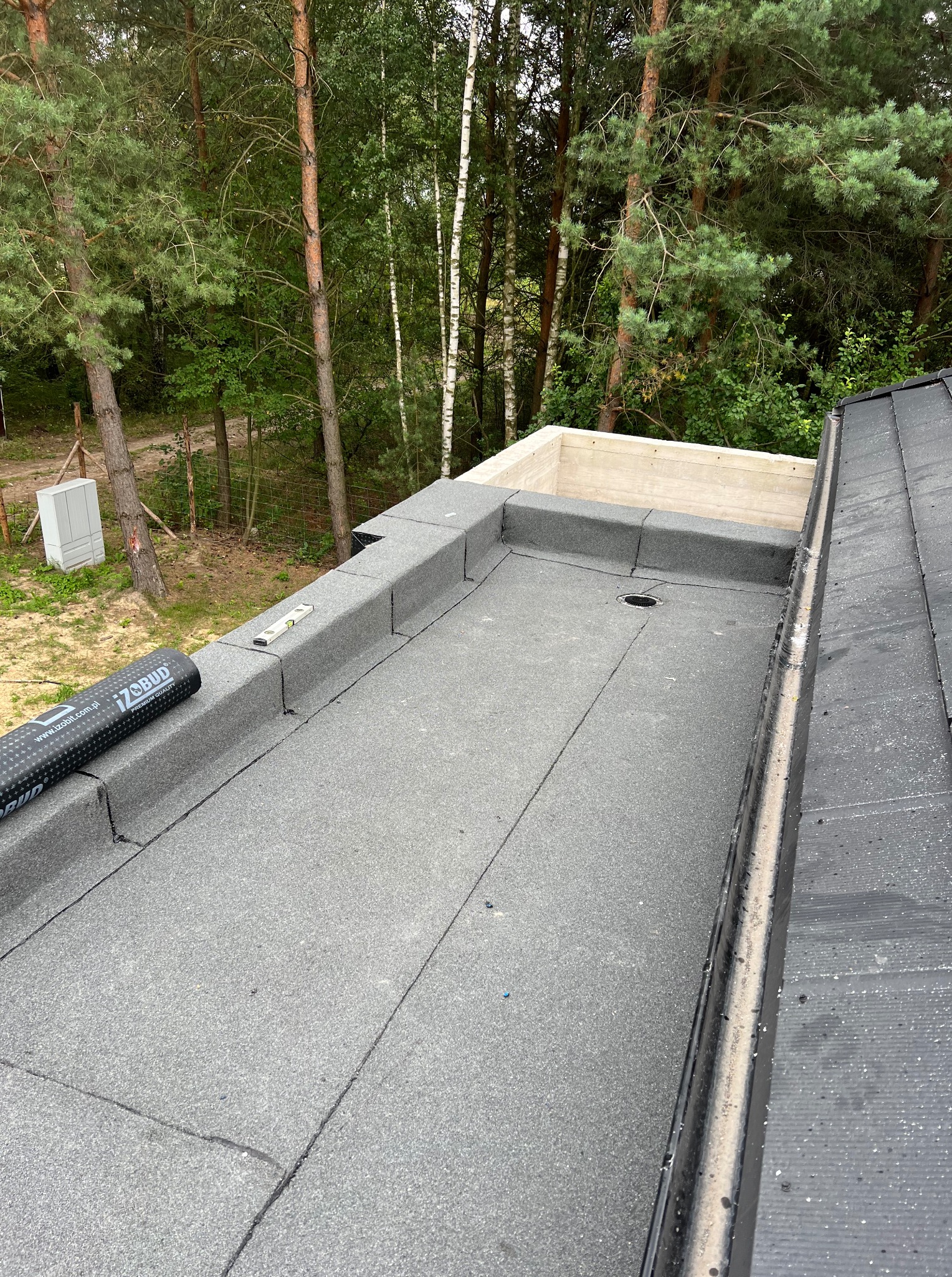 Płaski dach pokryty papą, z widocznym świetlikiem i rolką papy Izobud. Na dachu poziomica. W tle drzewa. Detal wykonania dachu.