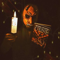 Brak - Osoba w masce demona trzymająca świecę i książkę 'Cienie Auroria' w mrocznym otoczeniu z dekoracyjnymi lampkami, tworząca niepokojącą atmosferę.