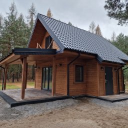 FIRMA HANDLOWO USŁUGOWA DREWTREND RADOSŁAW PIERZ - Gotowy dom szkieletowy z ciemnym dachem i drewnianą elewacją, otoczony lasem. Widoczna weranda z drewnianymi słupami i czarną podbitką dachu.
