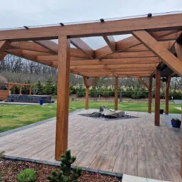 Drewniana pergola z przezroczystym zadaszeniem na tarasie z płytek, w tle trawnik i jacuzzi. Elementy dekoracyjne: kamienie, kwiaty w donicach.