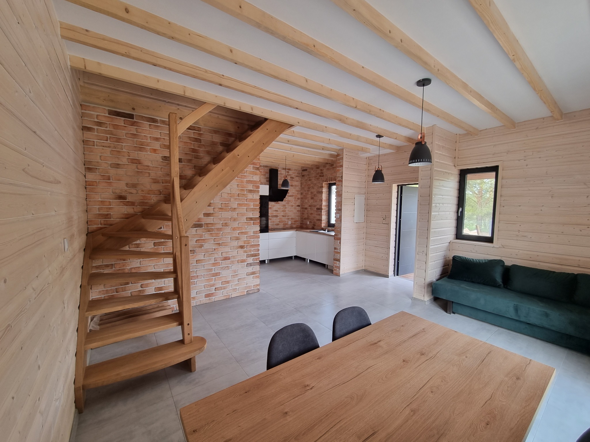 Jasne wnętrze domu szkieletowego: drewniane ściany, schody, ceglana ściana, widok na kuchnię, salon z sofą i drewniany stół. Nowoczesny design z naturalnymi materiałami.