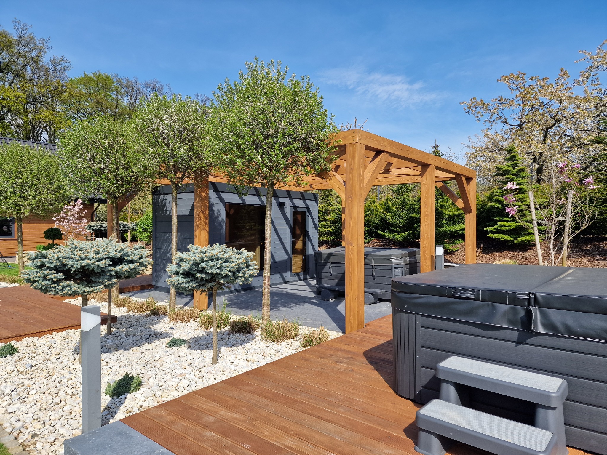 Drewniana pergola nad jacuzzi na tarasie z desek. W tle budynek z szarego drewna i drzewa. Całość w słoneczny dzień.