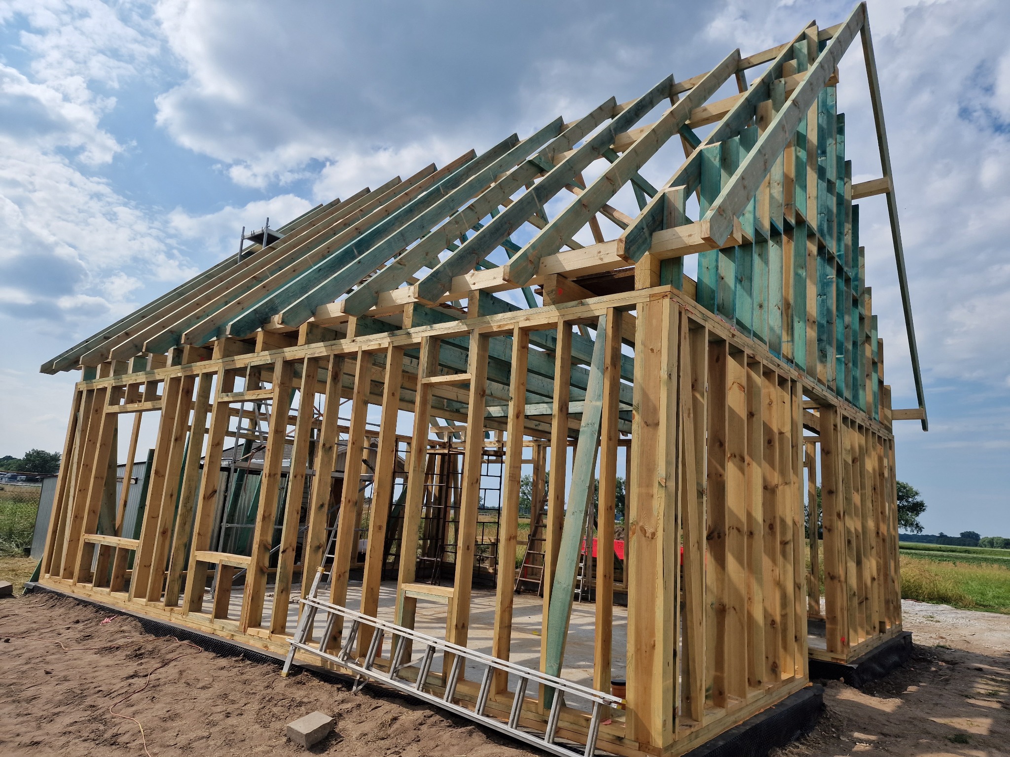 Konstrukcja szkieletowa domu w trakcie budowy, widoczne drewniane ramy ścian i dachu, drabina oparta o ścianę, błękitne niebo w tle. Prace budowlane w toku.