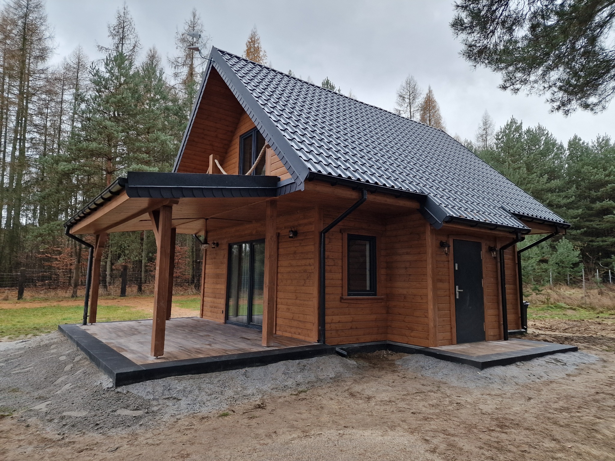 Gotowy dom szkieletowy z ciemnym dachem i drewnianą elewacją, otoczony lasem. Widoczna weranda z drewnianymi słupami i czarną podbitką dachu.