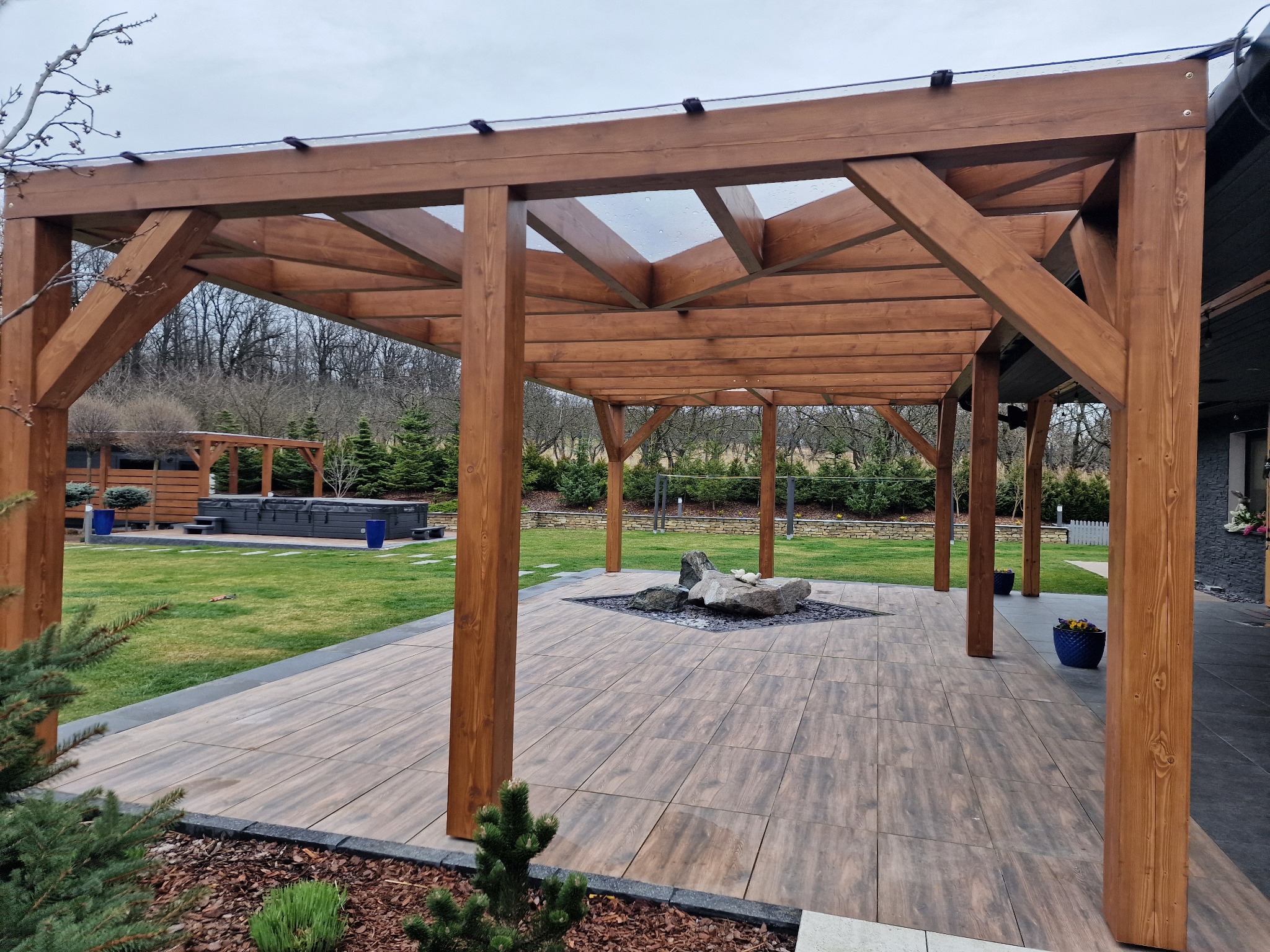 Drewniana pergola z przezroczystym zadaszeniem na tarasie z płytek, w tle trawnik i jacuzzi. Elementy dekoracyjne: kamienie, kwiaty w donicach.