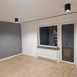 Firma Remontoo Budowlana - Wykończony pokój w Skalbmierzu: panele podłogowe, szara ściana z płyt, okno z grzejnikiem, czarne lampy sufitowe i listwa. Nowoczesne wnętrze po remoncie.