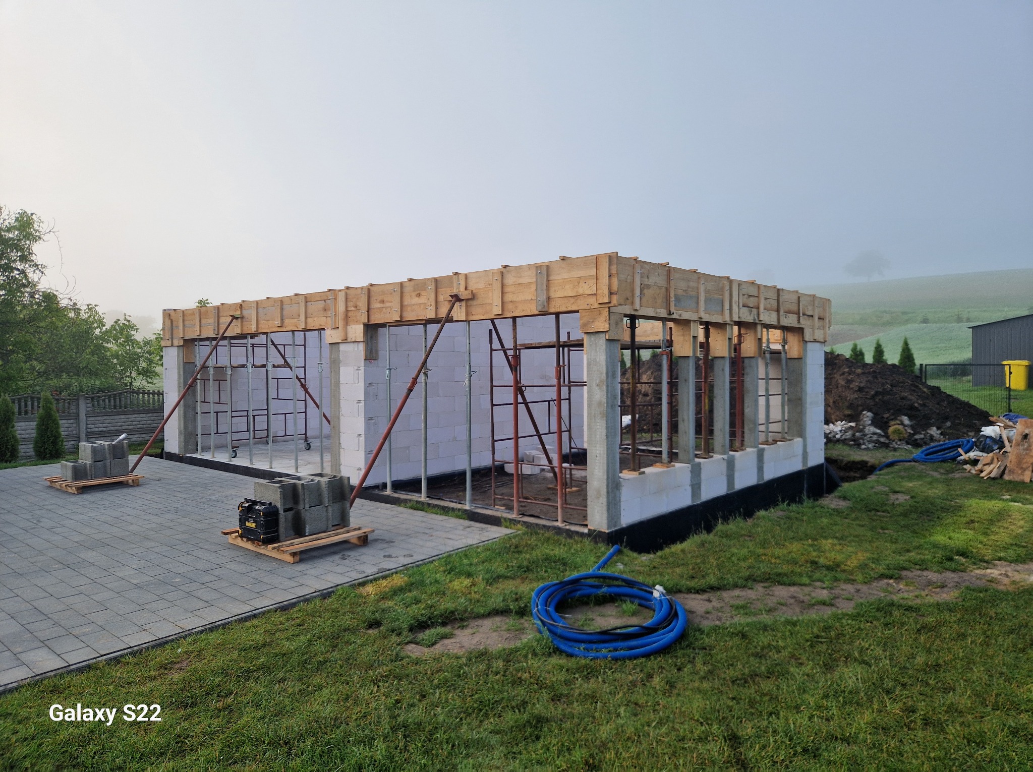 Budowa domu jednorodzinnego w trakcie realizacji: widoczne ściany z bloczków, szalunki na stropie i rusztowania. W tle zielony krajobraz.