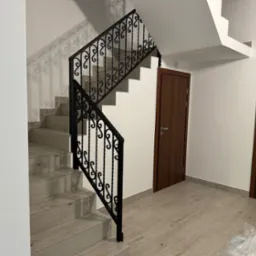 Klatka schodowa z czarną, kutą balustradą o dekoracyjnych elementach. Schody wyłożone płytkami imitującymi drewno. Dwa brązowe drzwi w tle.