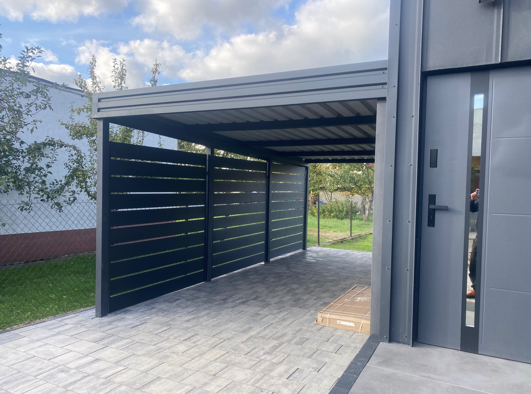 Nowoczesna, szara wiata garażowa z poziomymi panelami, wyłożona kostką brukową. W tle widoczny ogród i błękitne niebo z chmurami. Minimalistyczny design.