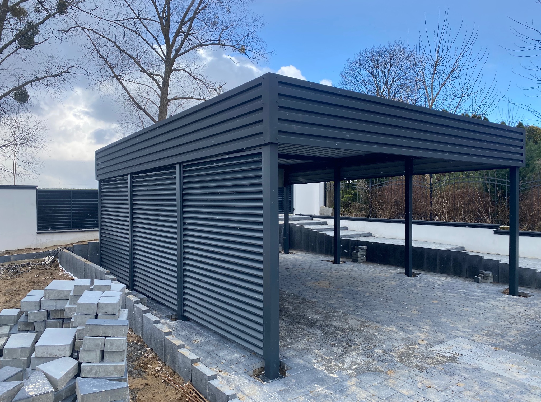 Nowoczesna, grafitowa wiata garażowa z poziomymi lamelami, kostka brukowa w trakcie układania, surowy krajobraz, błękitne niebo. Minimalistyczny design.