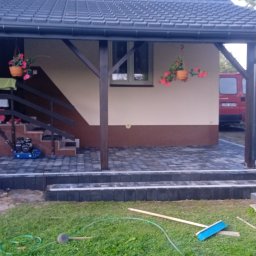 Łukasz Mlonek - Ułożona kostka brukowa w odcieniach szarości tworzy taras z schodami przed domem z drewnianą konstrukcją. Widoczna zagęszczarka i narzędzia brukarskie.