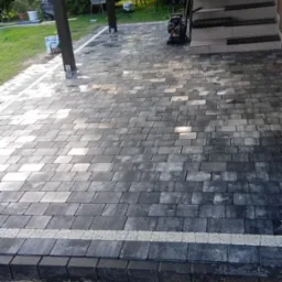 Ułożona, szara kostka brukowa z delikatnym wzorem, obrzeża z ciemnej kostki oddzielone białym pasem. W tle zielony ogród i fragment schodów.