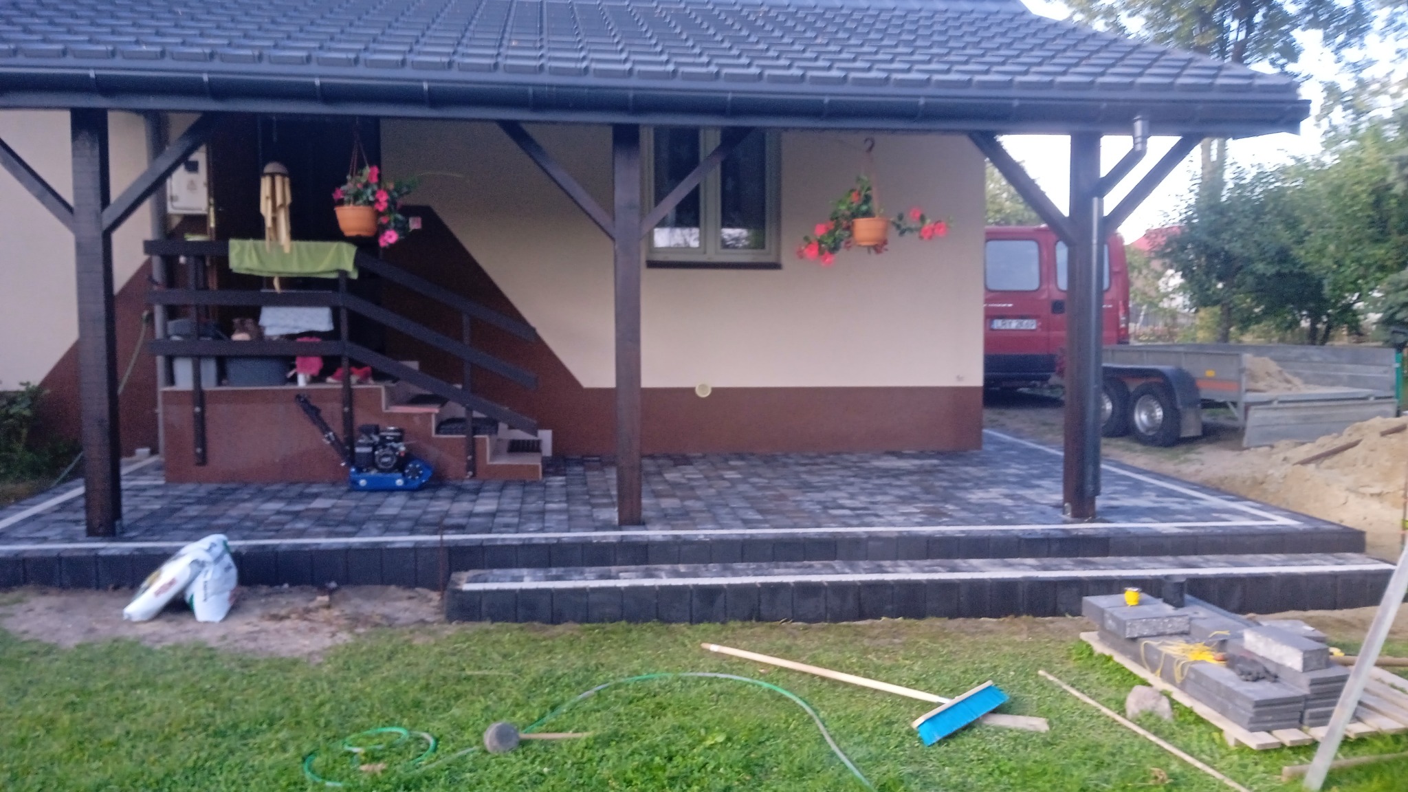 Ułożona kostka brukowa w odcieniach szarości tworzy taras z schodami przed domem z drewnianą konstrukcją. Widoczna zagęszczarka i narzędzia brukarskie.