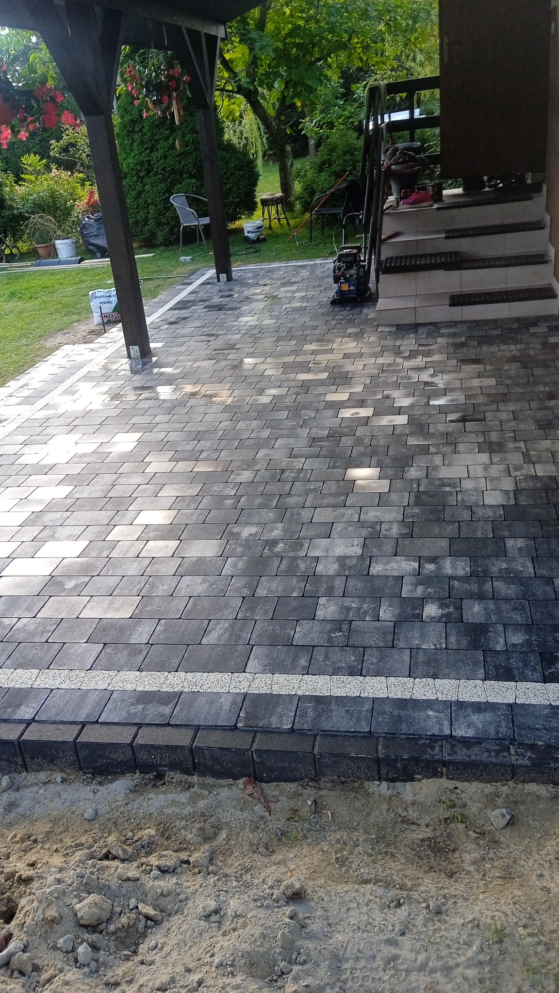 Ułożona, szara kostka brukowa z delikatnym wzorem, obrzeża z ciemnej kostki oddzielone białym pasem. W tle zielony ogród i fragment schodów.