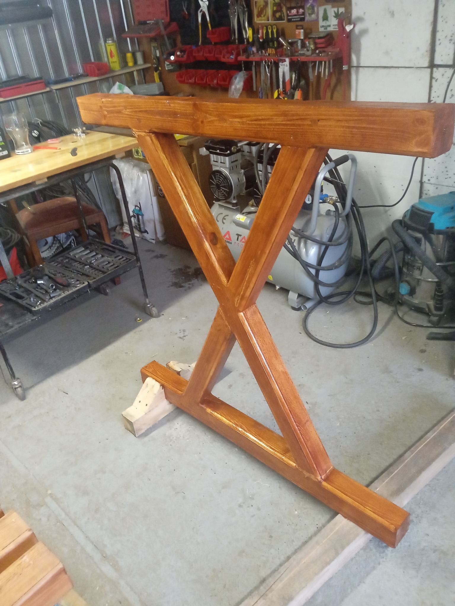 Drewniana podstawa stołu w kształcie litery X, pomalowana na brązowo, widoczna w warsztacie z narzędziami i sprzętem. Stojak na czas schnięcia podparty drewnianymi klockami.