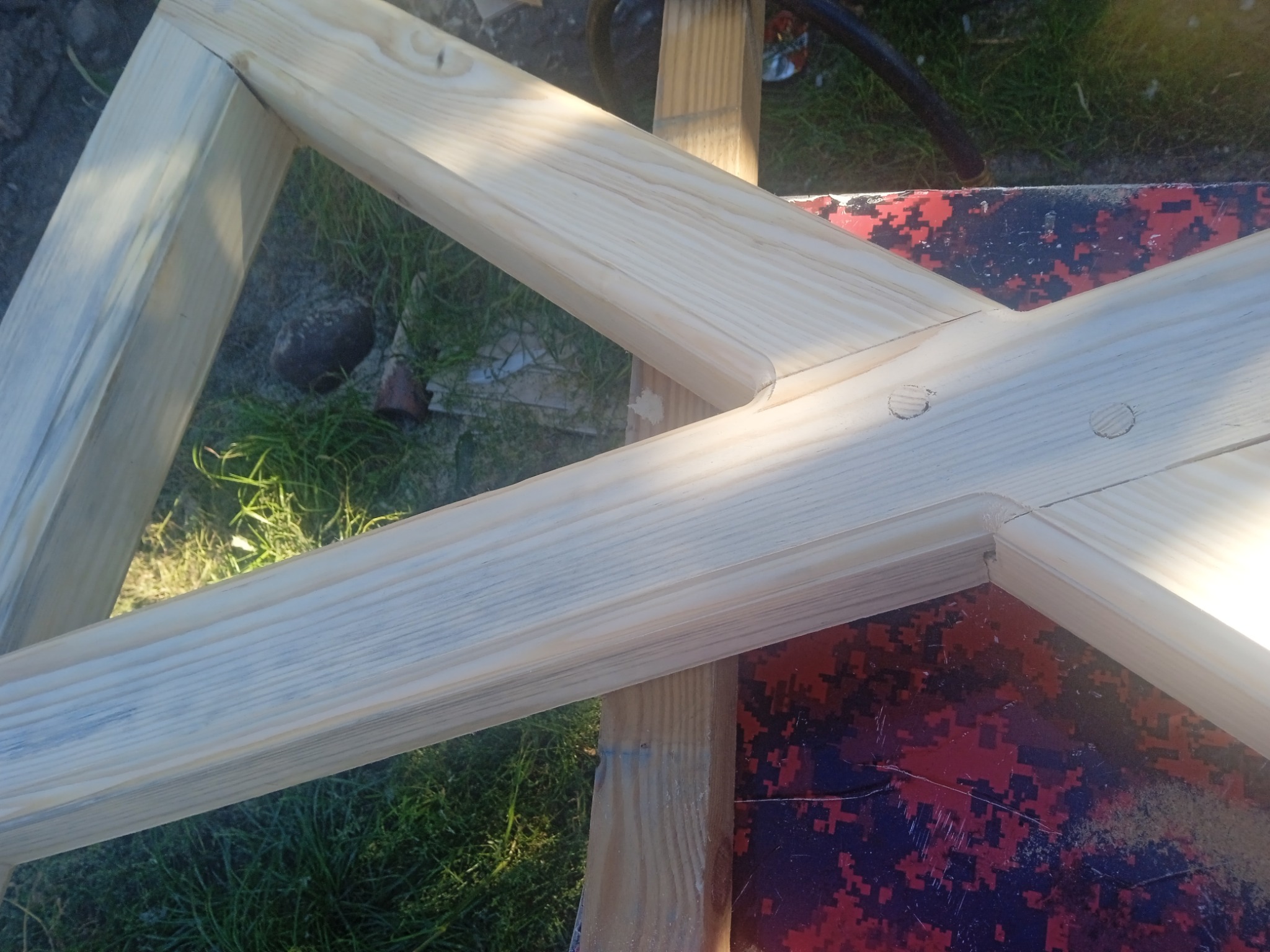 Fragment drewnianej konstrukcji ogrodzenia z jasnego drewna, z widocznymi połączeniami elementów, na tle trawy i deski z wzorem kamuflażu.