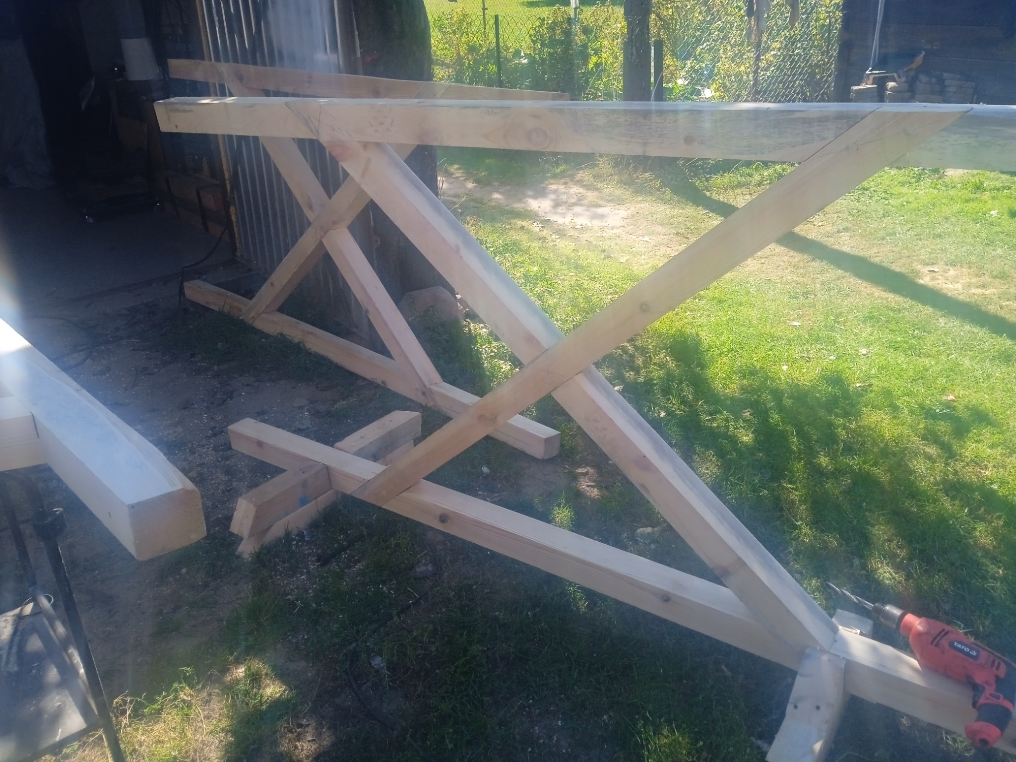 Konstrukcja drewniana w trakcie montażu na trawie, widoczna wiertarka. Elementy połączone pod kątem, tworzące szkielet. Praca w toku, jasne oświetlenie.