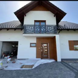 Elewacja domu jednorodzinnego po malowaniu, z widoczną nową stolarką drzwiową i okienną. Na pierwszym planie zabezpieczona folią kostka brukowa i farby.