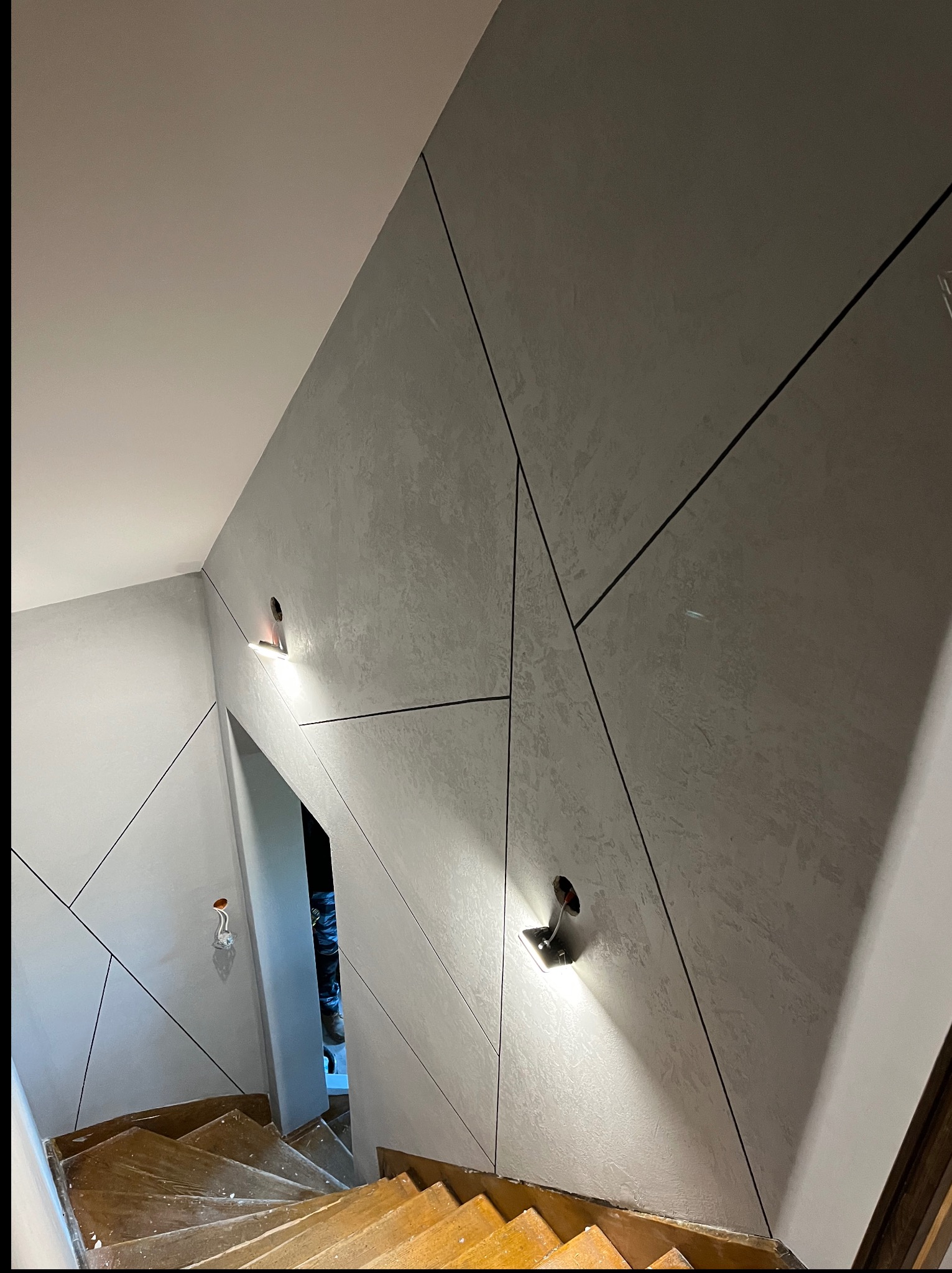 Szara ściana z geometrycznym wzorem i oświetleniem LED przy schodach. Widoczne niedokończone elementy instalacji elektrycznej. Nowoczesne wykończenie wnętrza.