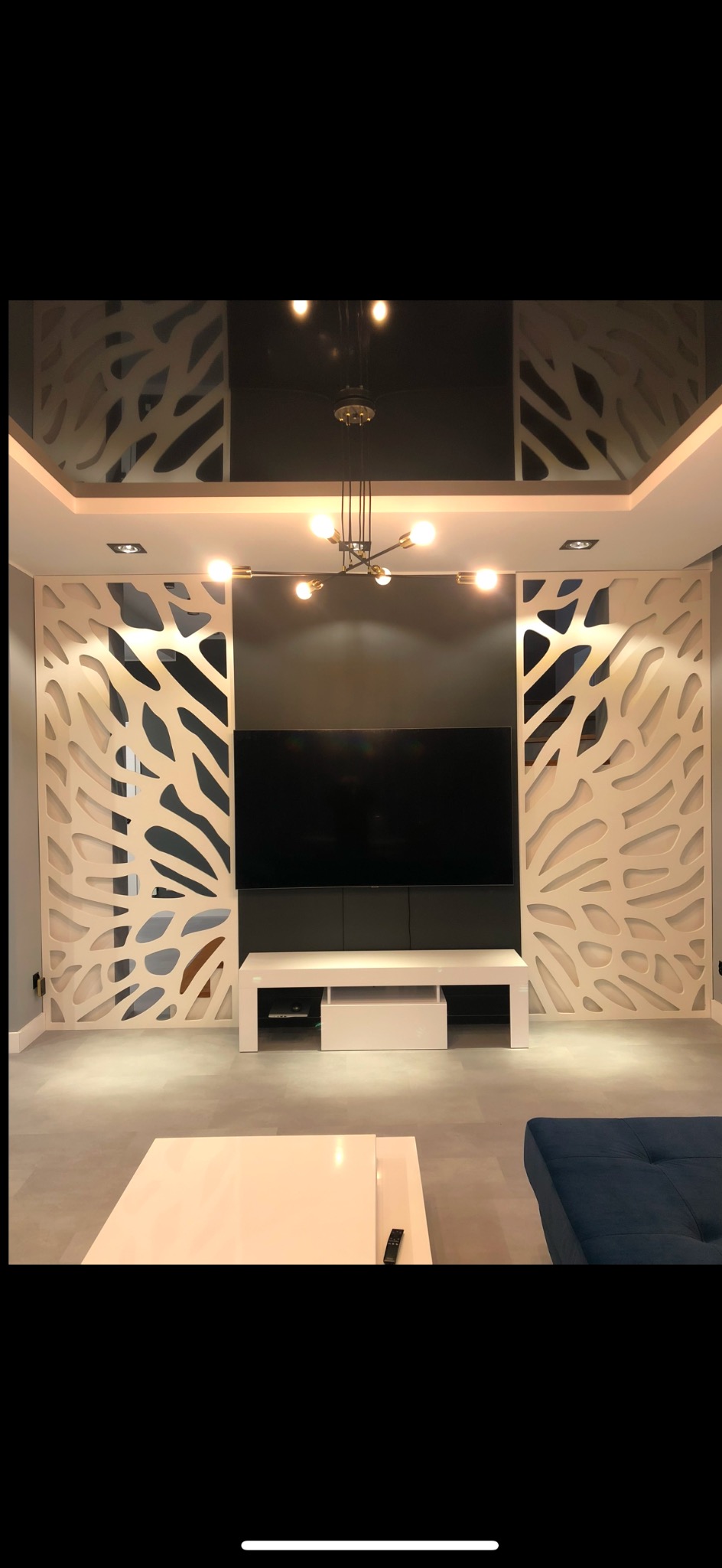 Nowoczesny salon z dekoracyjnymi panelami ażurowymi po bokach telewizora, podwieszany sufit z oświetleniem punktowym i designerska lampa wisząca.