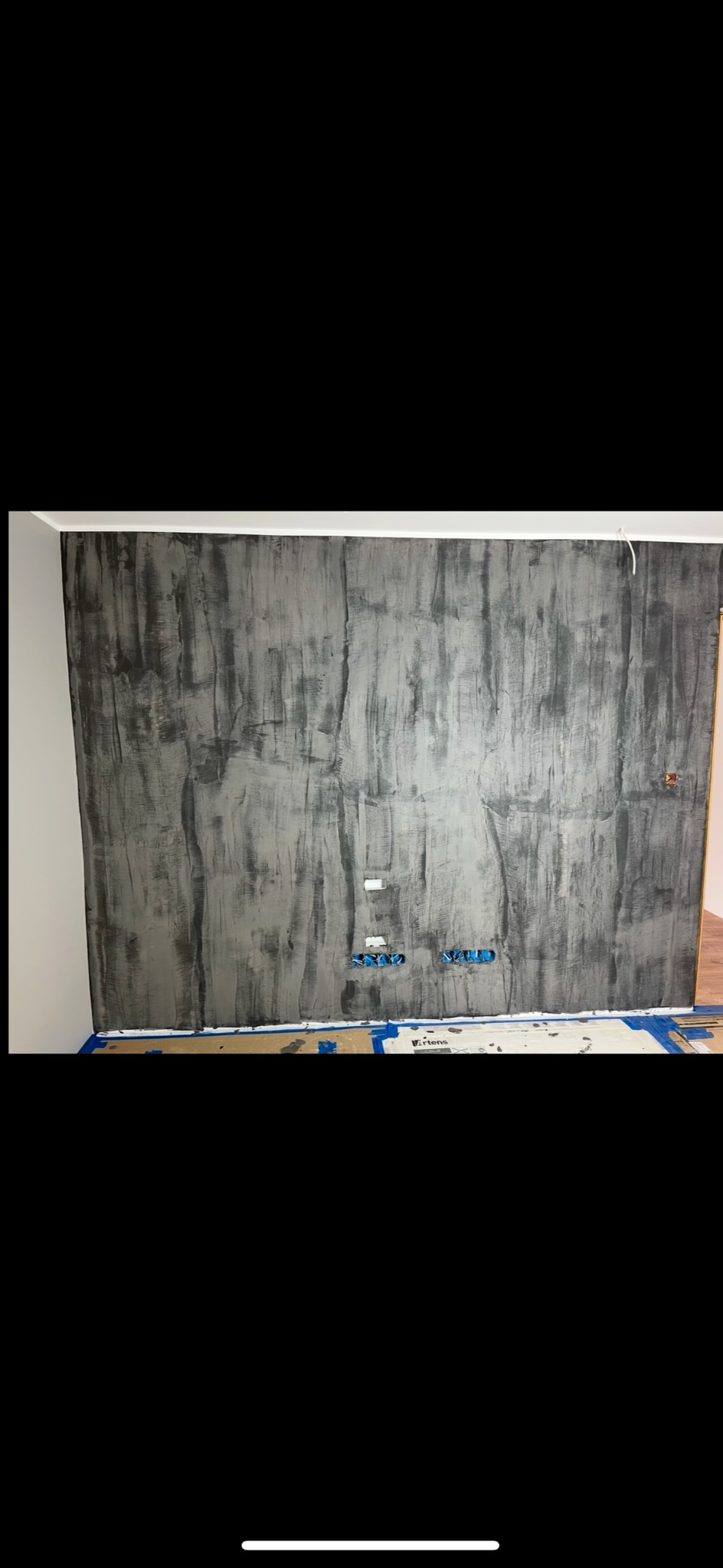 Ściana z efektem dekoracyjnym w odcieniach szarości, zabezpieczona taśmą malarską przy podłodze. Widoczne gniazdka elektryczne i fragment drewnianej podłogi.