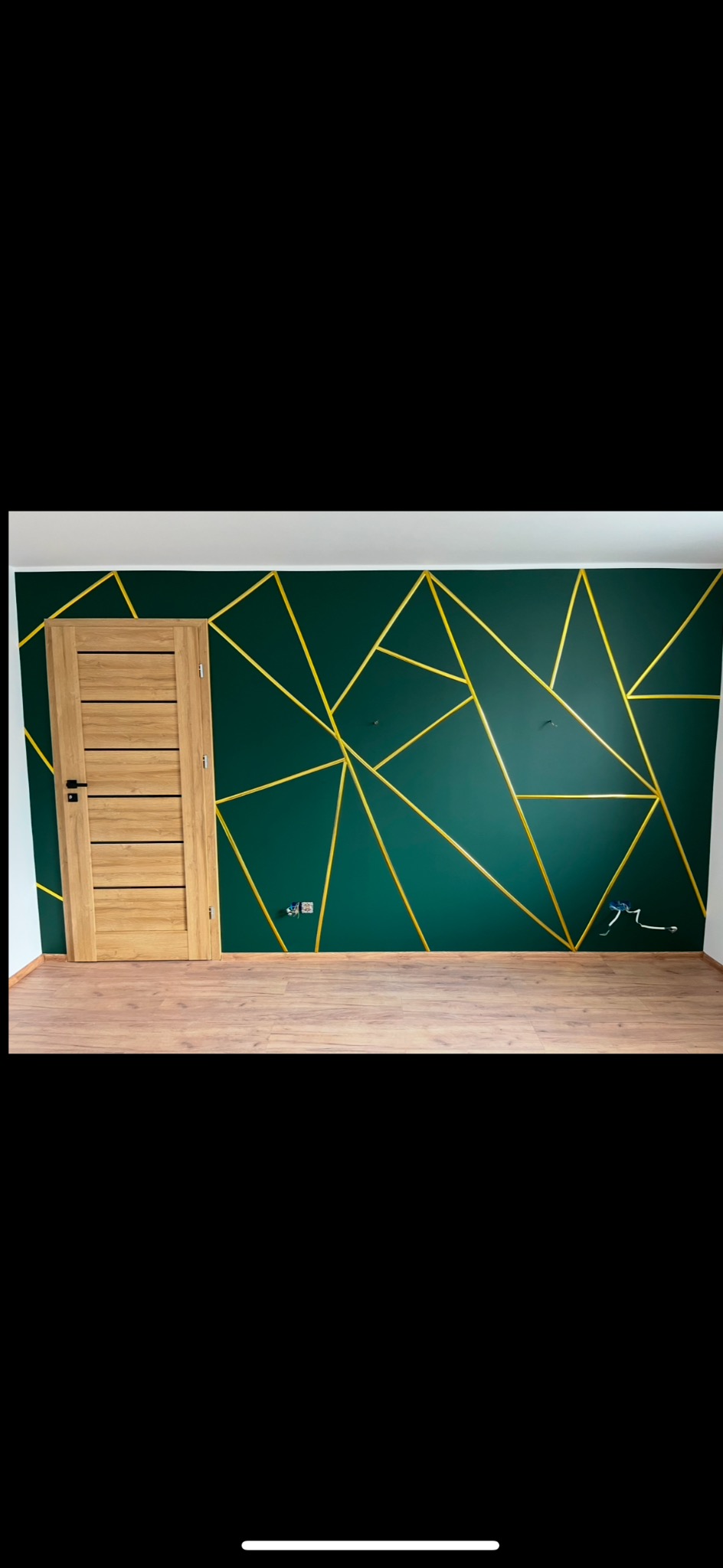 Zakończone wykończenie pokoju z zieloną ścianą, geometrycznym wzorem z żółtych pasków i drewnianą podłogą. Widoczne gniazdka elektryczne i zamontowane drzwi.